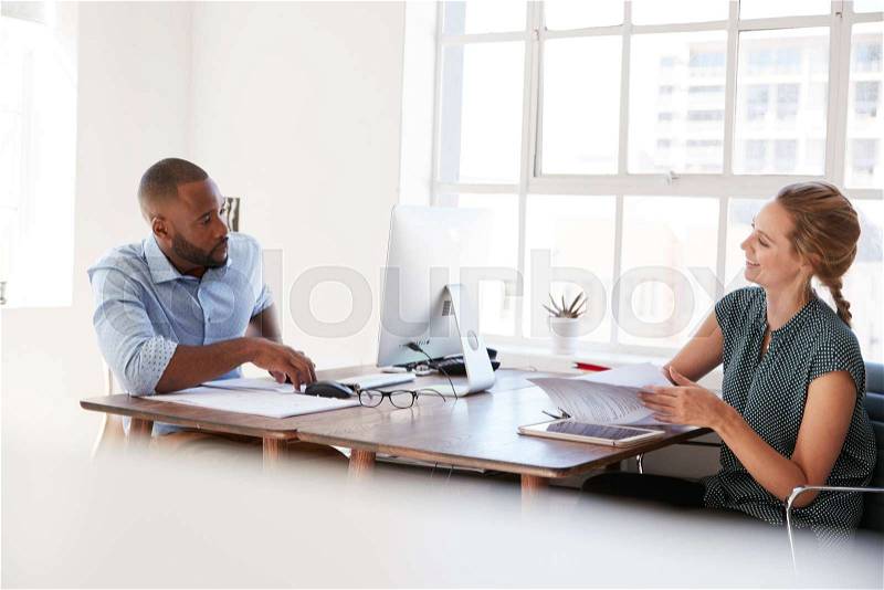 Young man and woman talking across their desks in an office, stock photo