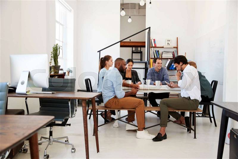 Creative team meet at a table in an office, one using phone, stock photo