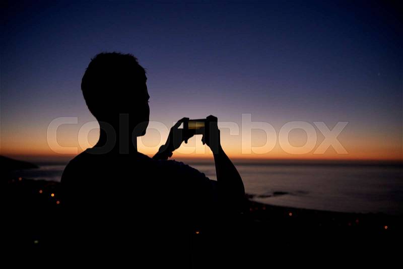 Man taking photos with phone on beach, silhouette at sunset, stock photo