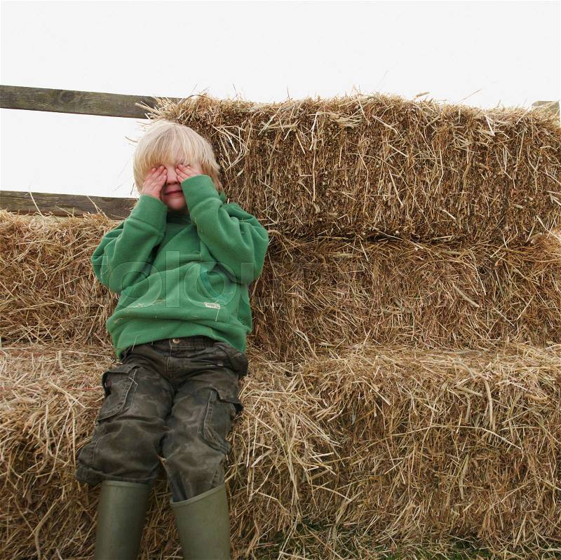Boy hiding eyes on hay bales, stock photo