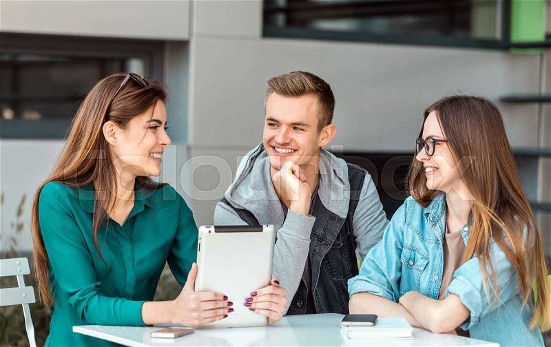Group of students wear glasses have work conversation outdoor cafe, table with tablet computer before modern building wall, sunny day, stock photo