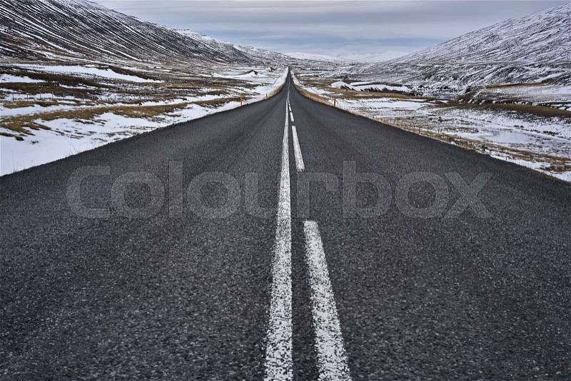Textured roadway with orange roadside pillars between the brown hills with remains of snow on the background of the cloudy sky in Iceland. Horizontal, stock photo
