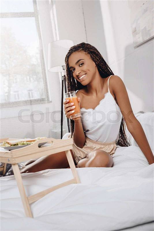 Happy african american woman with fresh juice and sandwich in bed, stock photo