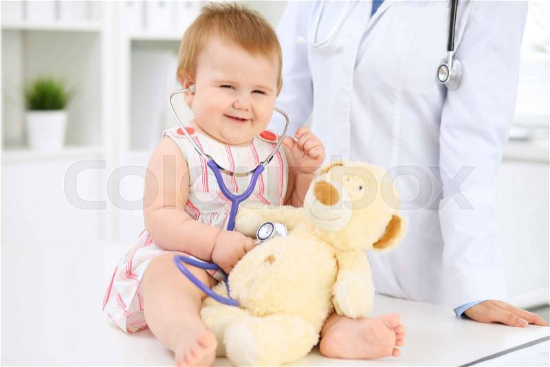 Happy cute baby at health exam at doctor\'s office. Toddler girl is sitting and keeping stethoscope and teddy bear, stock photo
