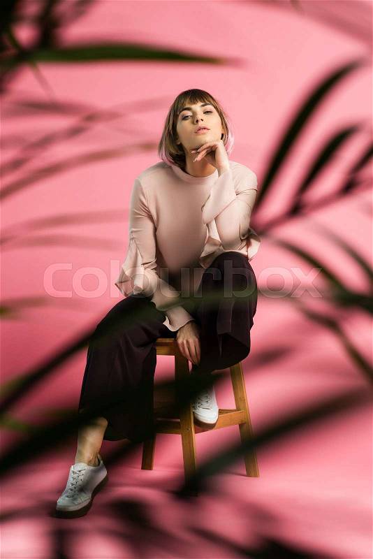 Young woman in trendy clothes posing on stool with hand on chin and looking at camera, stock photo