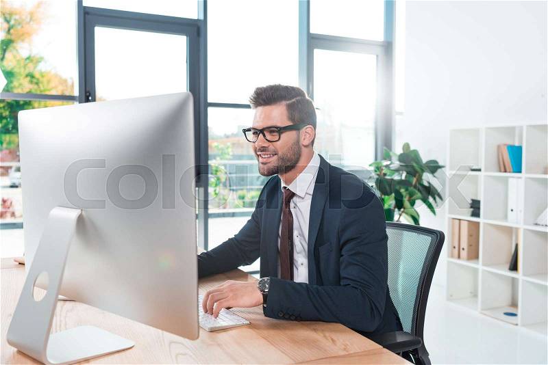 Smiling young businessman in eyeglasses using desktop computer in office, stock photo