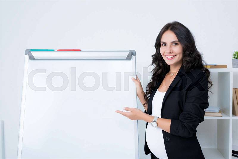 Happy pregnant businesswoman pointing at blank whiteboard and looking at camera, stock photo