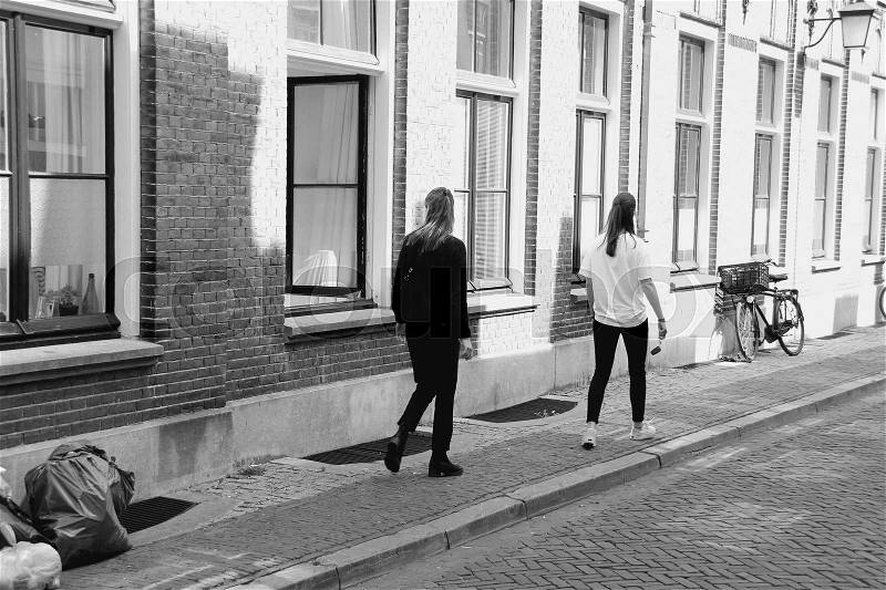 Two girlfriends are walking and one is looking inside the house through the open window in one of the streets in the residential area in the city Utrecht in the summer in black and white, stock photo