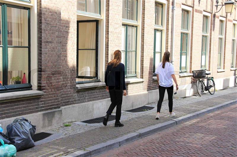 Two girlfriends are walking and one is looking inside the house through the open window in one of the streets in the residential area in the city Utrecht in the summer, stock photo
