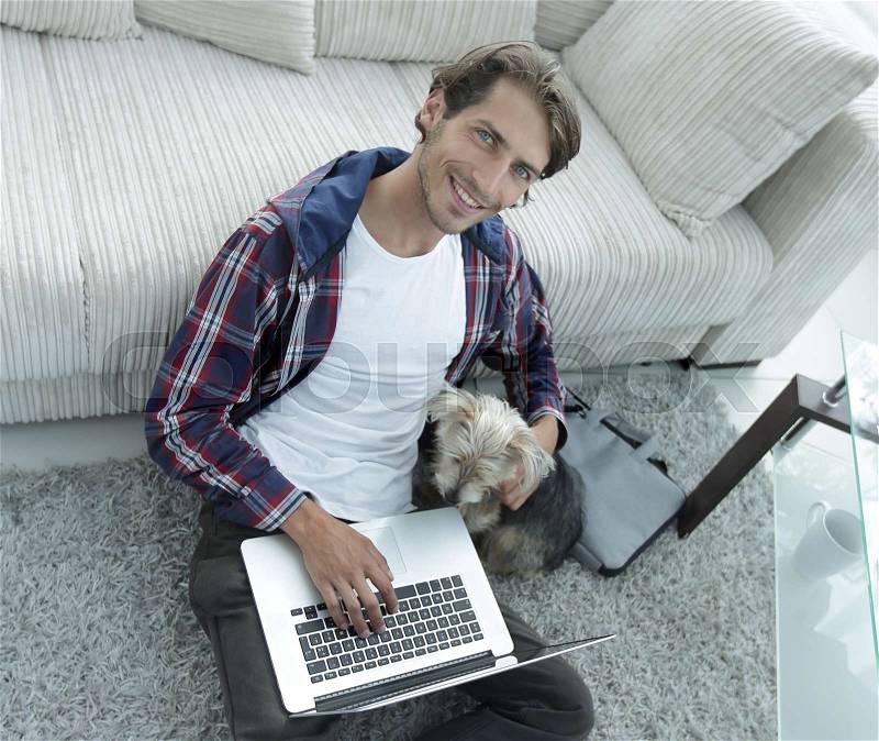 Handsome guy with laptop hugging his dog and sitting near the couch in a stylish living room, stock photo