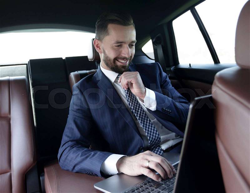 Smiling businessman reads information on laptop while sitting in car, stock photo