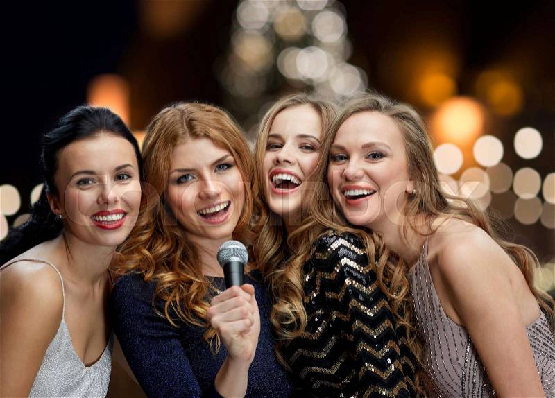 Holidays and people concept - happy women with microphone singing karaoke at new year party over christmas tree lights background, stock photo