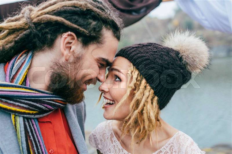 Stylish couple newlyweds smile and touching foreheads standing before a lake. Autumn wedding ceremony outdoors. Bride and groom with dreadlocks look at each other with tenderness and love. Close-up, stock photo