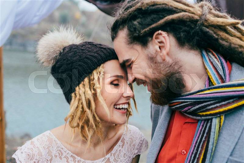 Stylish couple newlyweds smile and touching foreheads standing before a lake. Autumn wedding ceremony outdoors. Bride and groom with dreadlocks look at each other with tenderness and love. Close-up, stock photo