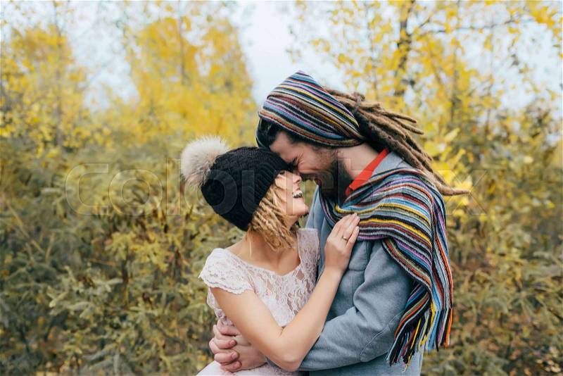 Bride in a knitted hat with a pom pom, hat covering her eyes. Groom in a colorful scarf. Happy young newlywed couple touching foreheads and noses in park. Close-up portrait, stock photo