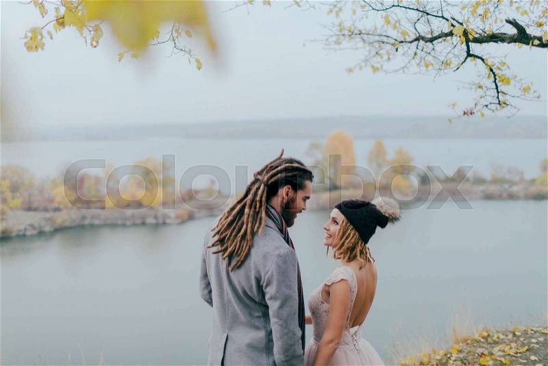 Attractive couple newlyweds laugh and smile. Autumn wedding ceremony outdoors. Stylish bride and groom with dreadlocks look at each other standing before a lake. Back view, stock photo
