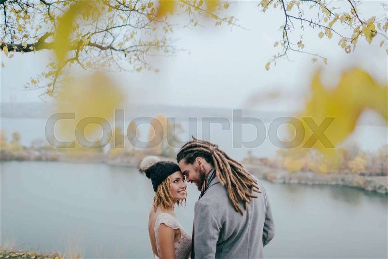 Stylish couple newlyweds smile and touching foreheads standing before a lake. Bride and groom with dreadlocks look at each other standing before a lake. Autumn wedding ceremony outdoors. Back view, stock photo