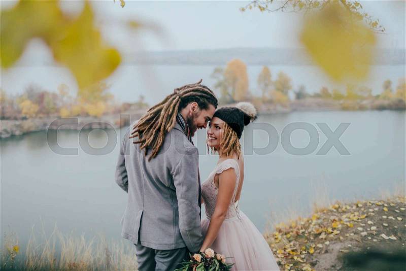 Stylish couple newlyweds smile and touching foreheads standing before a lake. Bride and groom with dreadlocks look at each other standing before a lake. Autumn wedding ceremony outdoors. Back view, stock photo