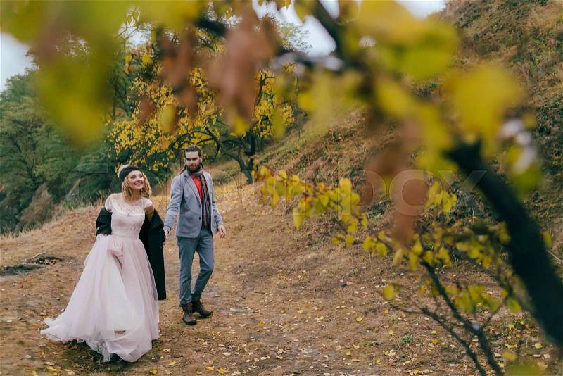 A happy couple is walking on a trail in an autumn forest. Bride and groom with dreadlocks are looking at each other on nature. Wedding ceremony outdoors. Full length portrait, stock photo