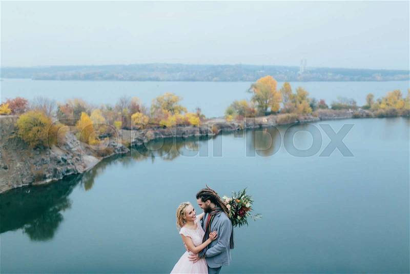 Stylish couple newlyweds smile and hugging standing before a lake. Bride and groom with dreadlocks look at each other. Autumn wedding ceremony outdoors. Overall view, stock photo