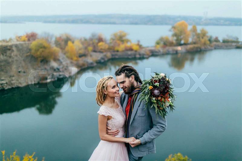 Stylish couple newlyweds smile and hugging standing before a lake. Bride and groom with dreadlocks look at each other. Autumn wedding ceremony outdoors. Close-up portrait, stock photo
