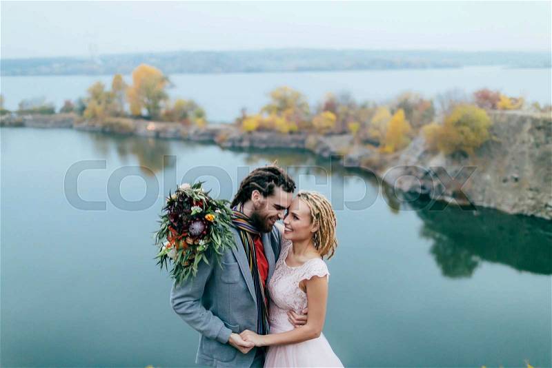 Stylish couple newlyweds smile and hugging standing before a lake. Bride and groom with dreadlocks look at each other. Autumn wedding ceremony outdoors. Close-up portrait, stock photo