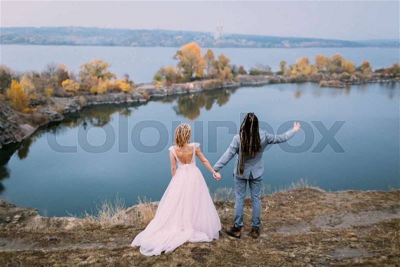 Back view of stylish couple newlyweds with hands up are posing before a lake on the hill. Autumn wedding ceremony outdoors. Full length portrait, stock photo
