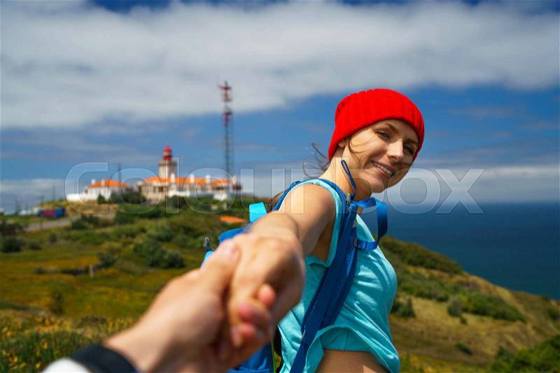 Follow me - happy young woman in a red hat and with a backpack behind her back pulling guy\'s hand. Hand in hand walking to the lighthouse. Concept of carefree modern life, stock photo