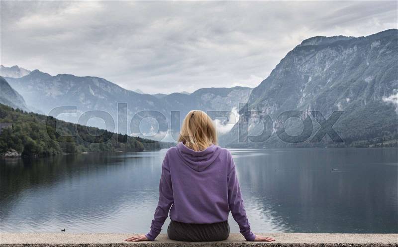 Solitary woman wearing purple hoodie watching tranquil overcast morning scene at lake Bohinj, Alps mountains, Slovenia, stock photo