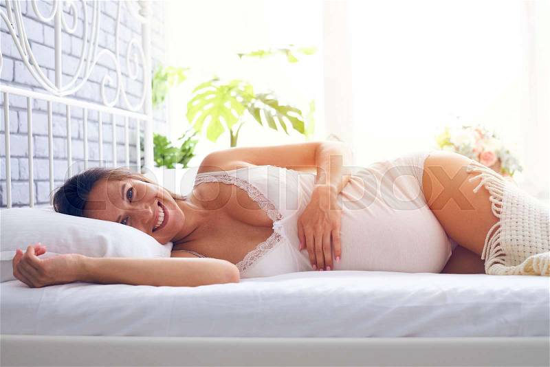 Closeup of smiling pregnant woman lying on side on the bed, holding hand on pregnant belly , stock photo