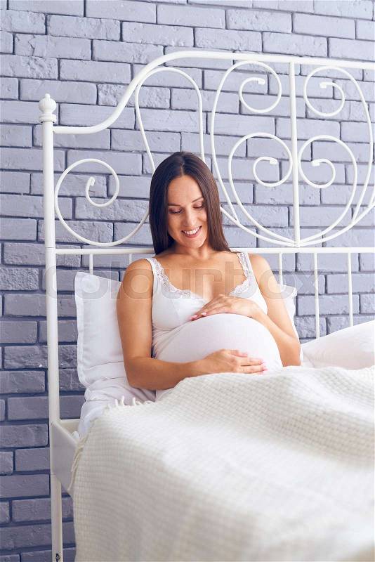 Vertical of elated brunette looking at pregnant belly on bed , stock photo