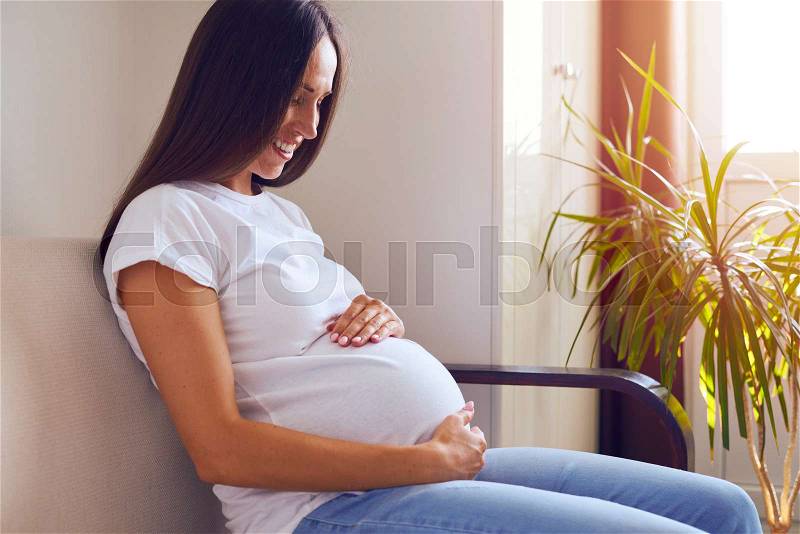 Side view of beautiful pregnant woman sitting on sofa at home, stock photo