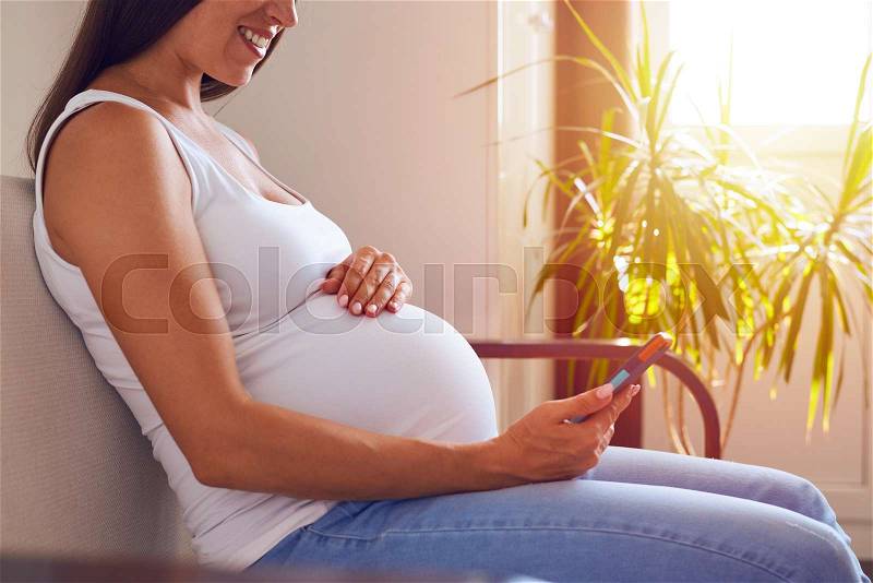 Cropped shot of mother-to-be sitting on sofa and using phone, stock photo