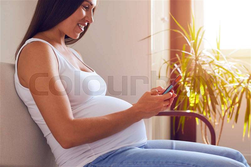 Side view of happy mother-to-be sitting on sofa and using phone, stock photo
