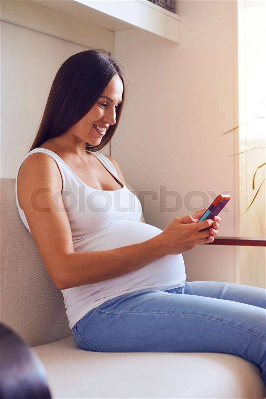 Side view of pregnant woman browsing in smartphone on sofa, stock photo