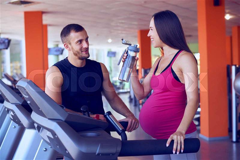 Closeup of pregnant woman drinking water while speaking to personal trainer in gym, stock photo