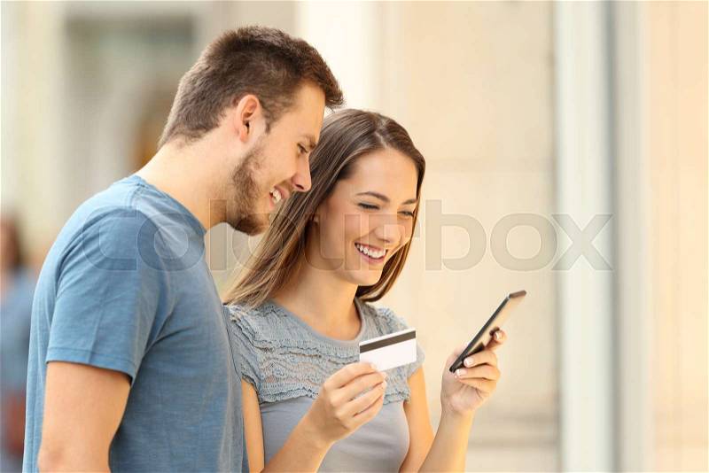 Shoppers buying on line with credit card and smart phone standing beside a storefront on the street, stock photo