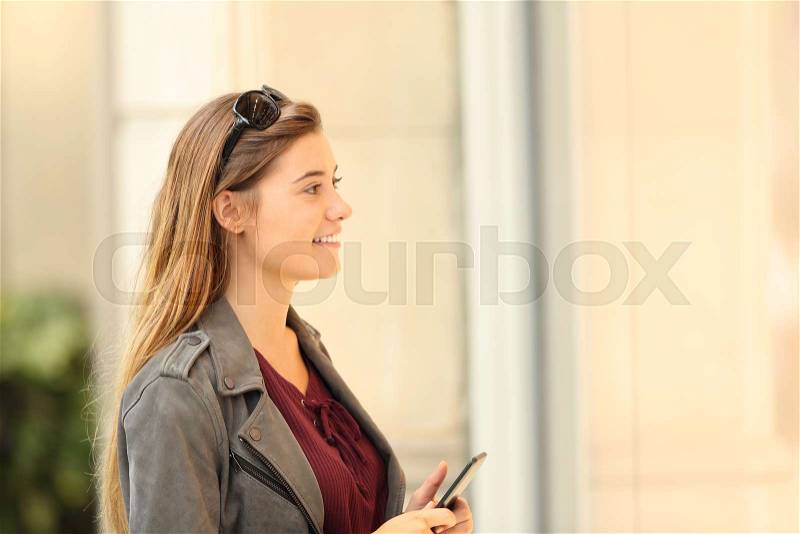 Side view of a happy shopper watching a storefront holding a smart phone outdoor on the street, stock photo