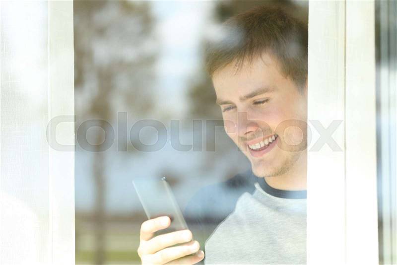 View through a window of a happy man using a smart phone at home, stock photo