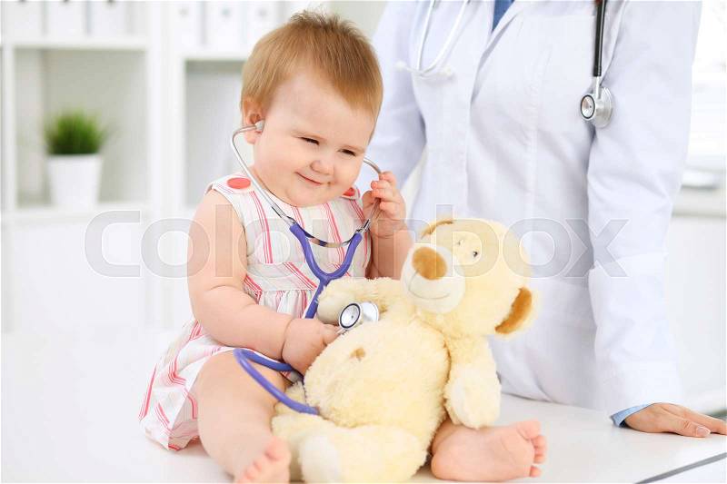 Happy cute baby at health exam at doctor's office. Toddler girl is sitting and keeping stethoscope and teddy bear, stock photo