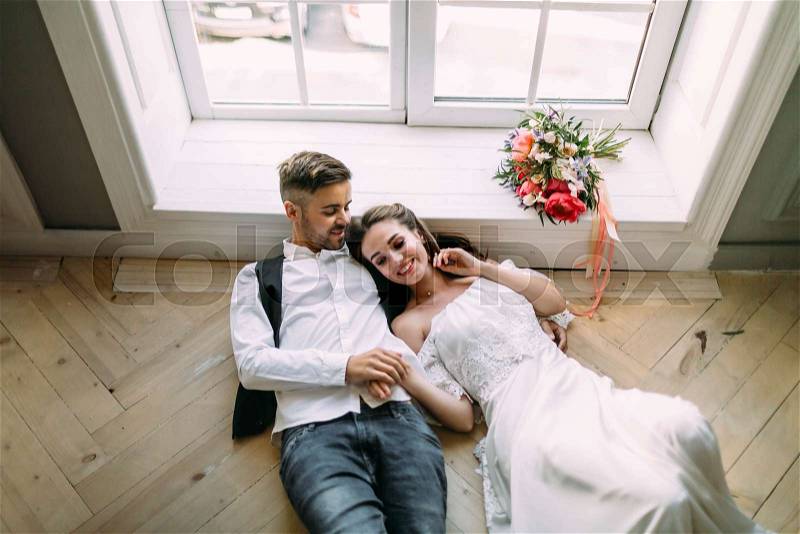 Cheerful bride and groom holding hands and lie on the wooden floor near the panoramic window. Wedding morning. Top view, stock photo