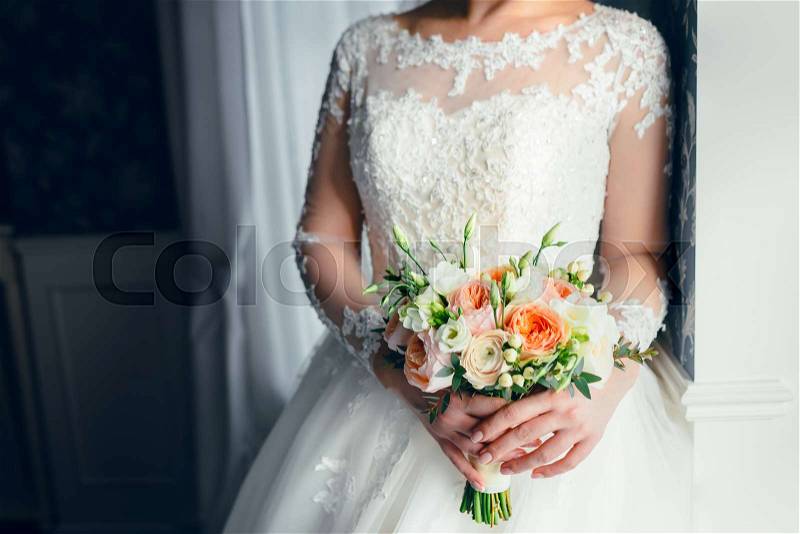 A beautiful bride is standing near the window and holding a wedding bouquet with white roses and peach peonies. Close-up, indoor, stock photo