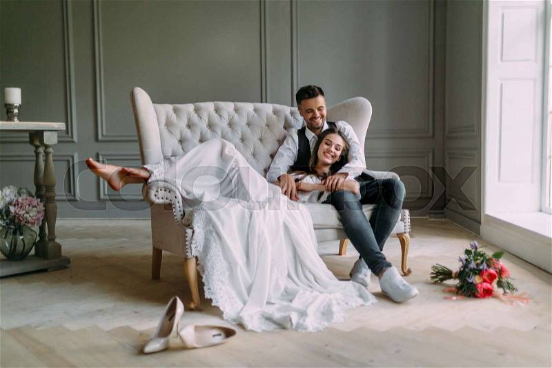 Young bride in tender lace dress lies on knees of a handsome groom in blue jeans. Cheerful newlyweds are laughing. Full length. Indoors, stock photo