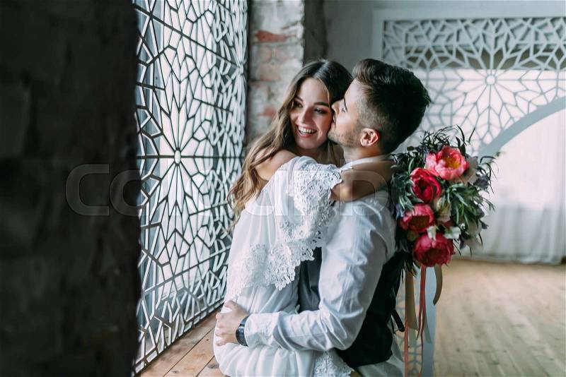 Cheerful young bride hugging groom while he kisses her in cheek and embraces her waist on the windowsill. Close-up, stock photo