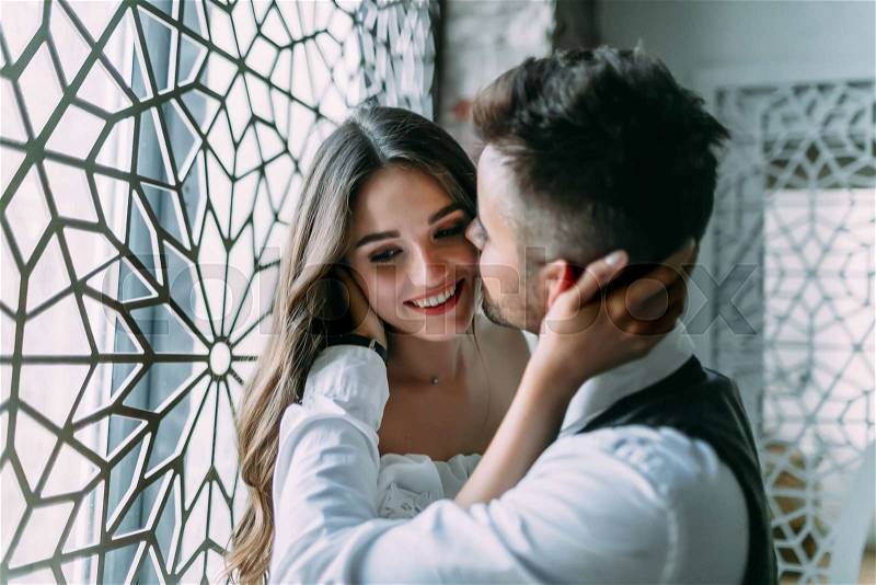 Cheerful young bride embraces groom\'s head while he kisses her in cheek on the vintage window background. Close-up, stock photo