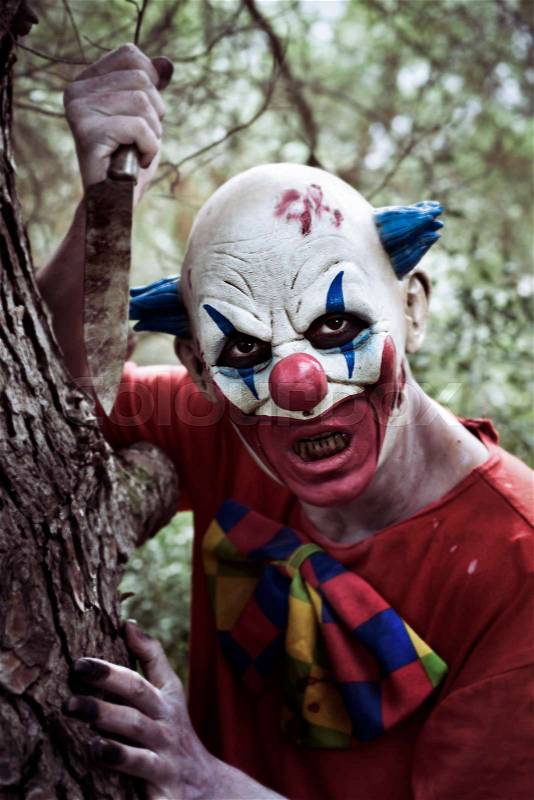 Closeup of a scary evil clown wearing a dirty and ragged red costume, and wielding a big knife in the woods, stock photo