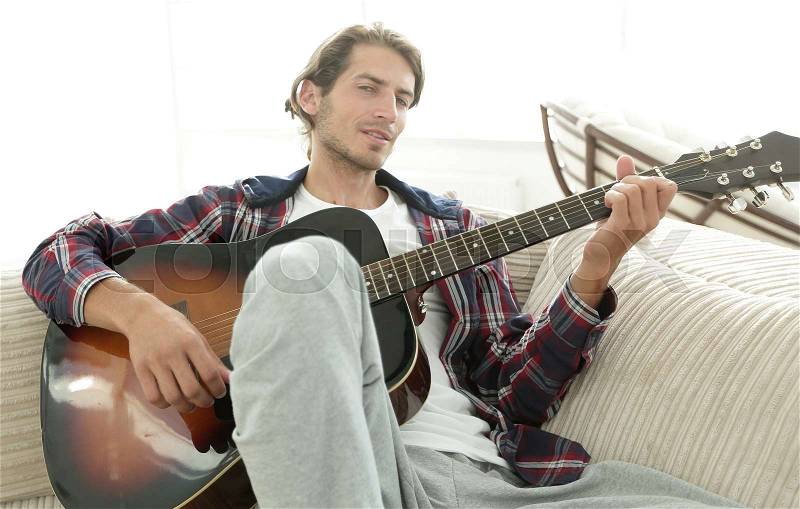 Stylish guy with guitar sitting on sofa in living room, stock photo