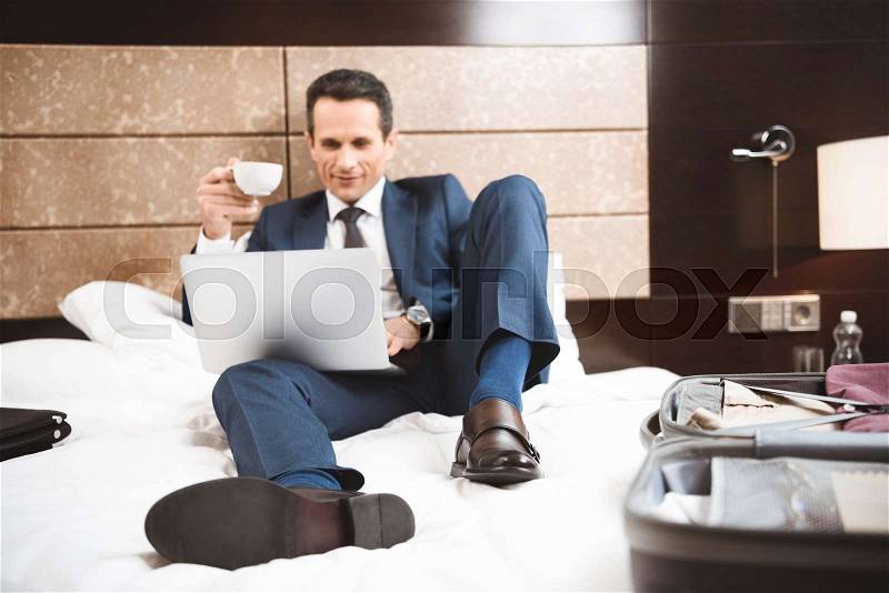 Young businessman in formal suit using his laptop while sitting on bed in hotel room, stock photo
