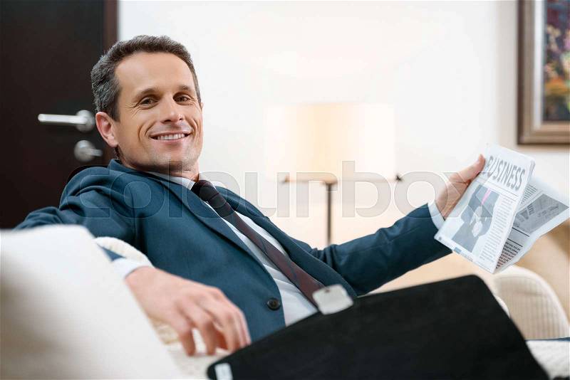 Smiling businessman in formal suit sitting in armchair with newspaper , stock photo