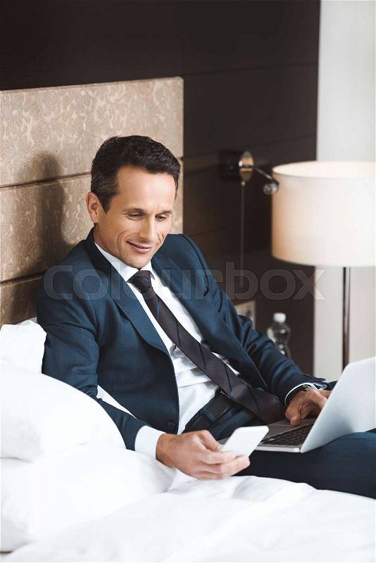 Smiling businessman in formal suit sitting on hotel room bed and using his smartphone and laptop, stock photo
